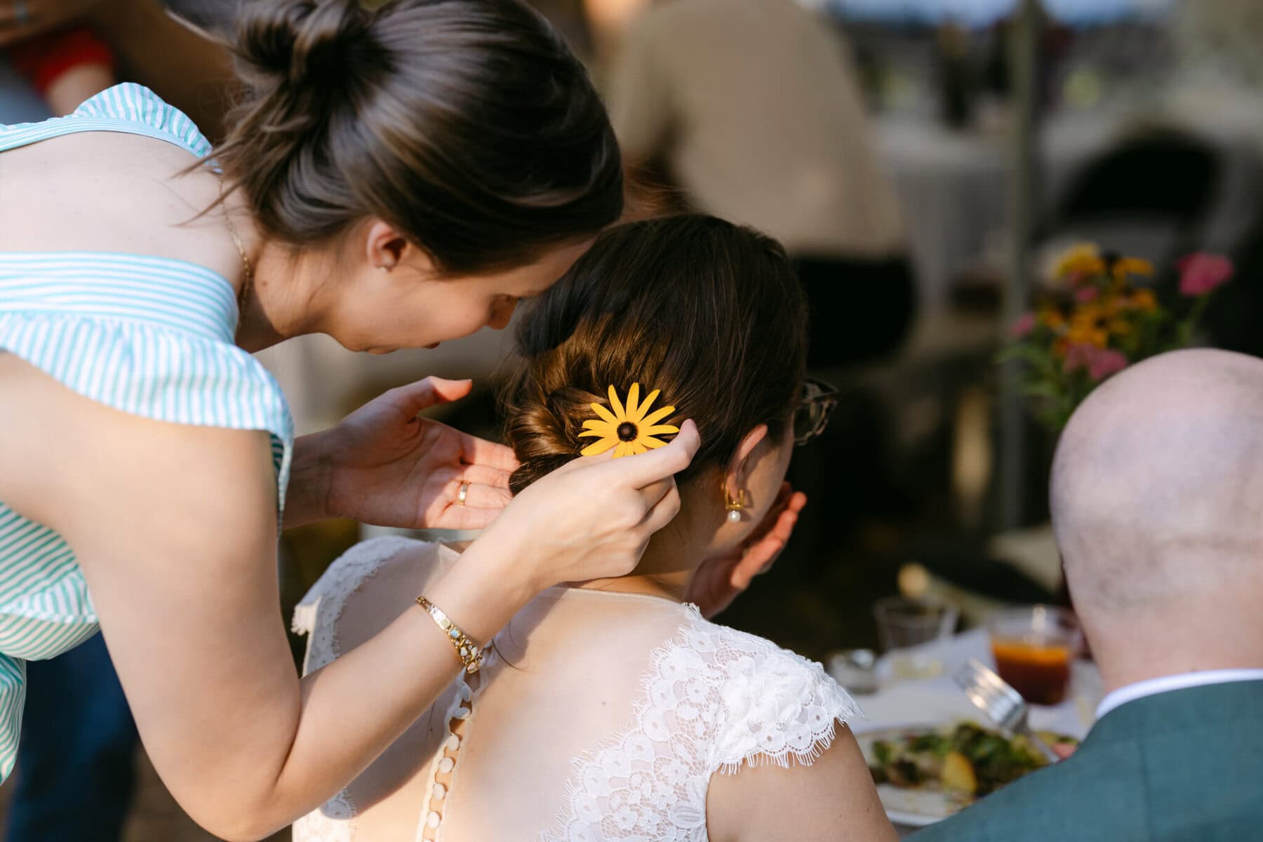 A Worcester Wedding at Cook Pond 79 At a Worcester Wedding by Cook Pond, a woman adorns another’s hair with a yellow flower as guests mingle near a table of food.