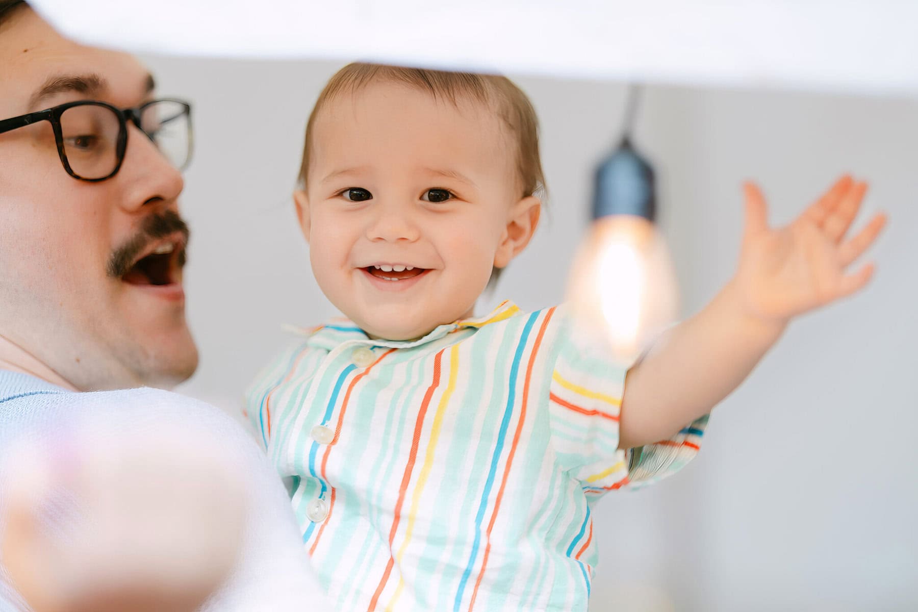 A Worcester Wedding at Cook Pond 80 An adult holding a smiling baby in a colorful striped shirt, arm raised, with a light bulb in the background at a Worcester Wedding.