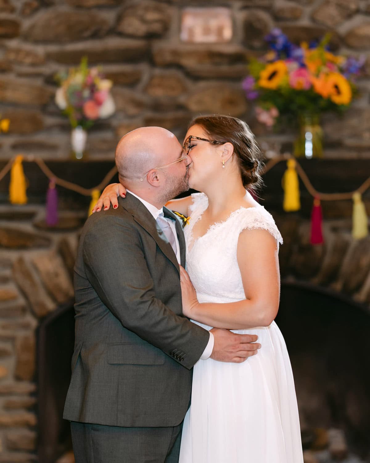 A Worcester Wedding at Cook Pond 82 A bride and groom kiss in front of a flower-adorned stone fireplace at their beautiful Worcester wedding.