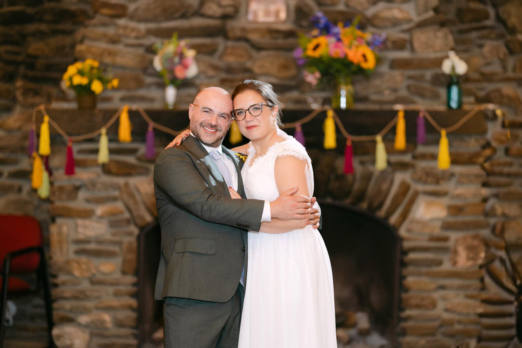 A Worcester Wedding at Cook Pond 83 A bride and groom stand close together, smiling, at their Worcester Wedding in front of a stone fireplace adorned with flowers and tassels.