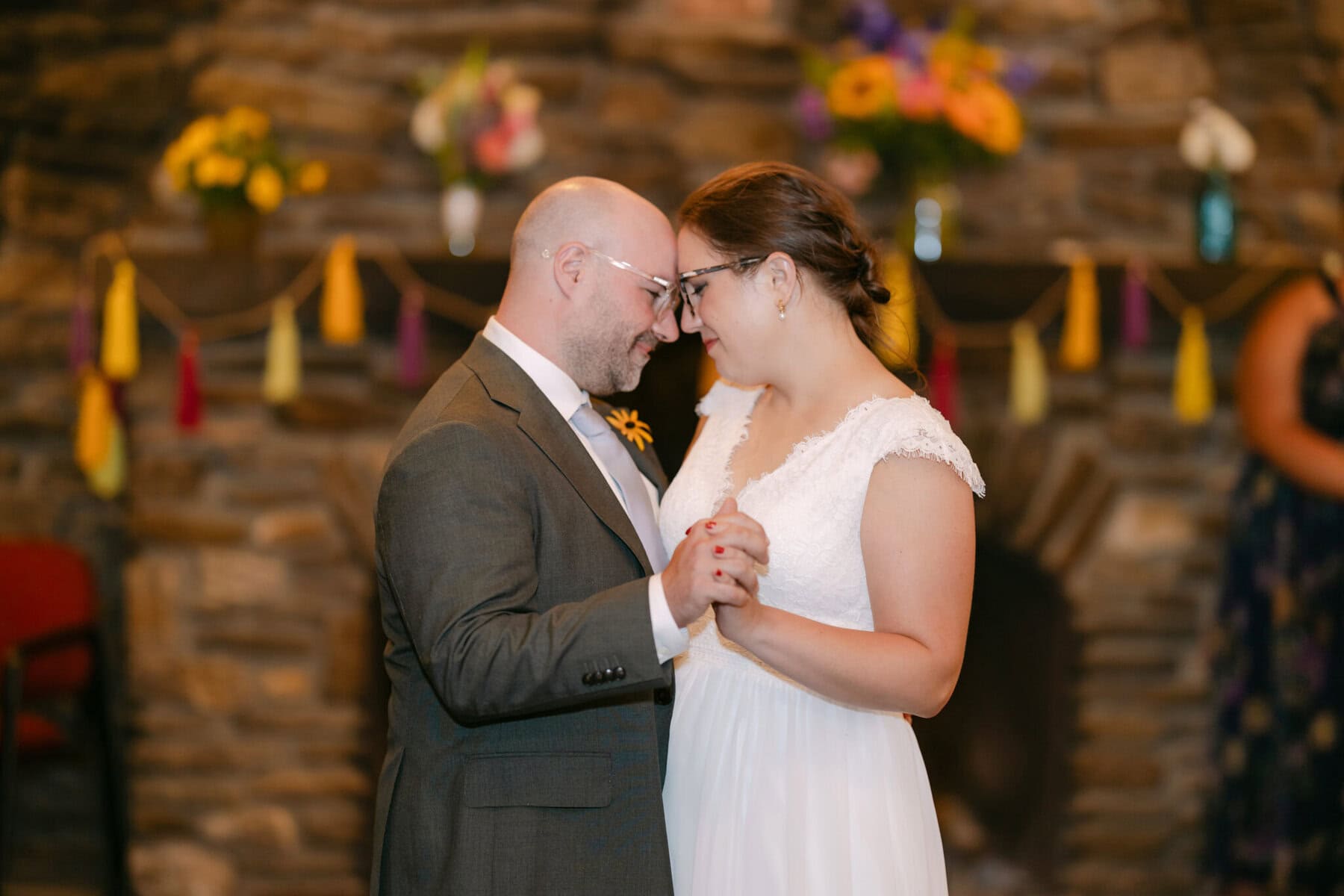 A Worcester Wedding at Cook Pond 85 A couple in formal attire dances closely indoors at a Worcester Wedding, framed by a stone wall adorned with flowers and colorful tassels.